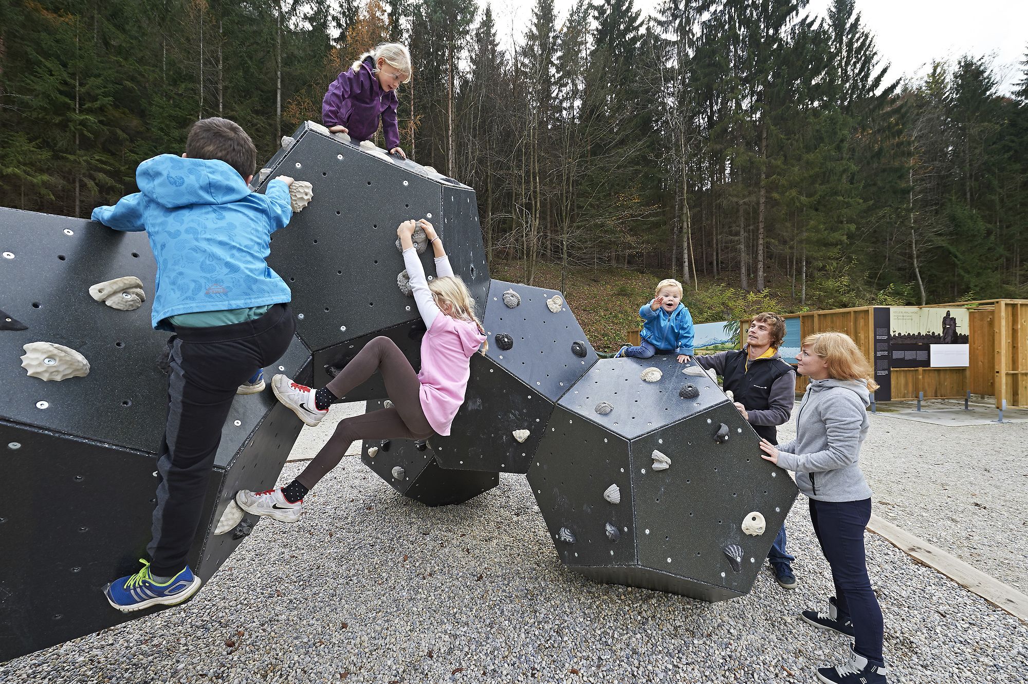 A group of children is climbing a modern, geometric climbing structure outdoors, surrounded by a forest.